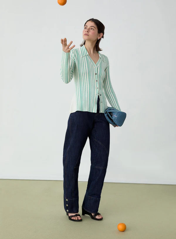 Woman wearing a green-and-white striped cardigan and dark wide-leg jeans, holding a blue handbag while tossing an orange in the air against a plain background.