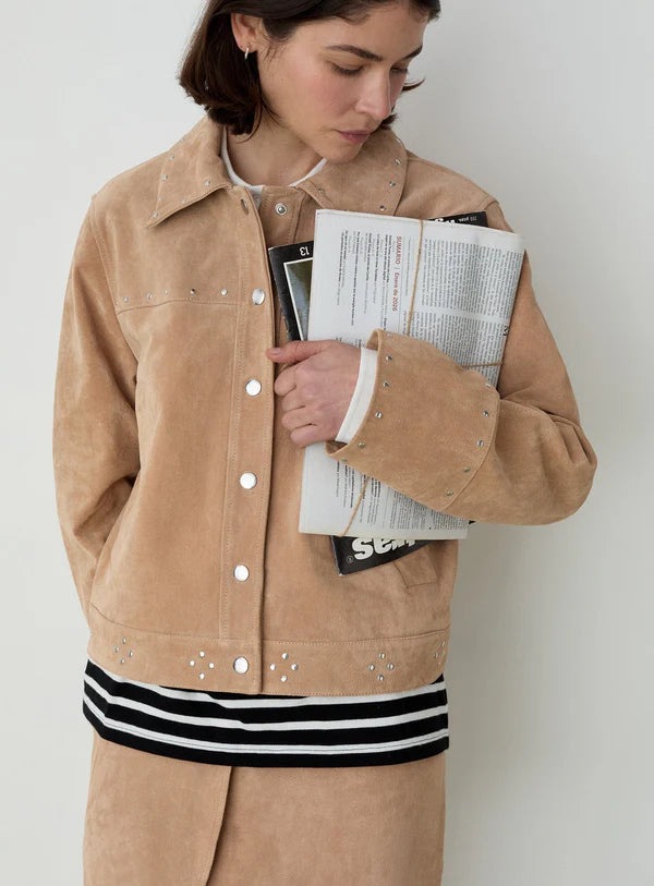 Woman wearing a tan studded jacket and matching skirt over a black-and-white striped top, holding magazines while looking down, against a plain background.