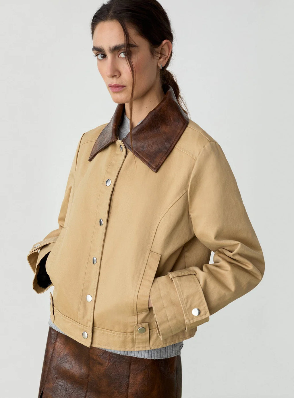 Woman wearing a tan jacket with a brown leather collar and brown skirt, posing with hands in pockets against a plain background.