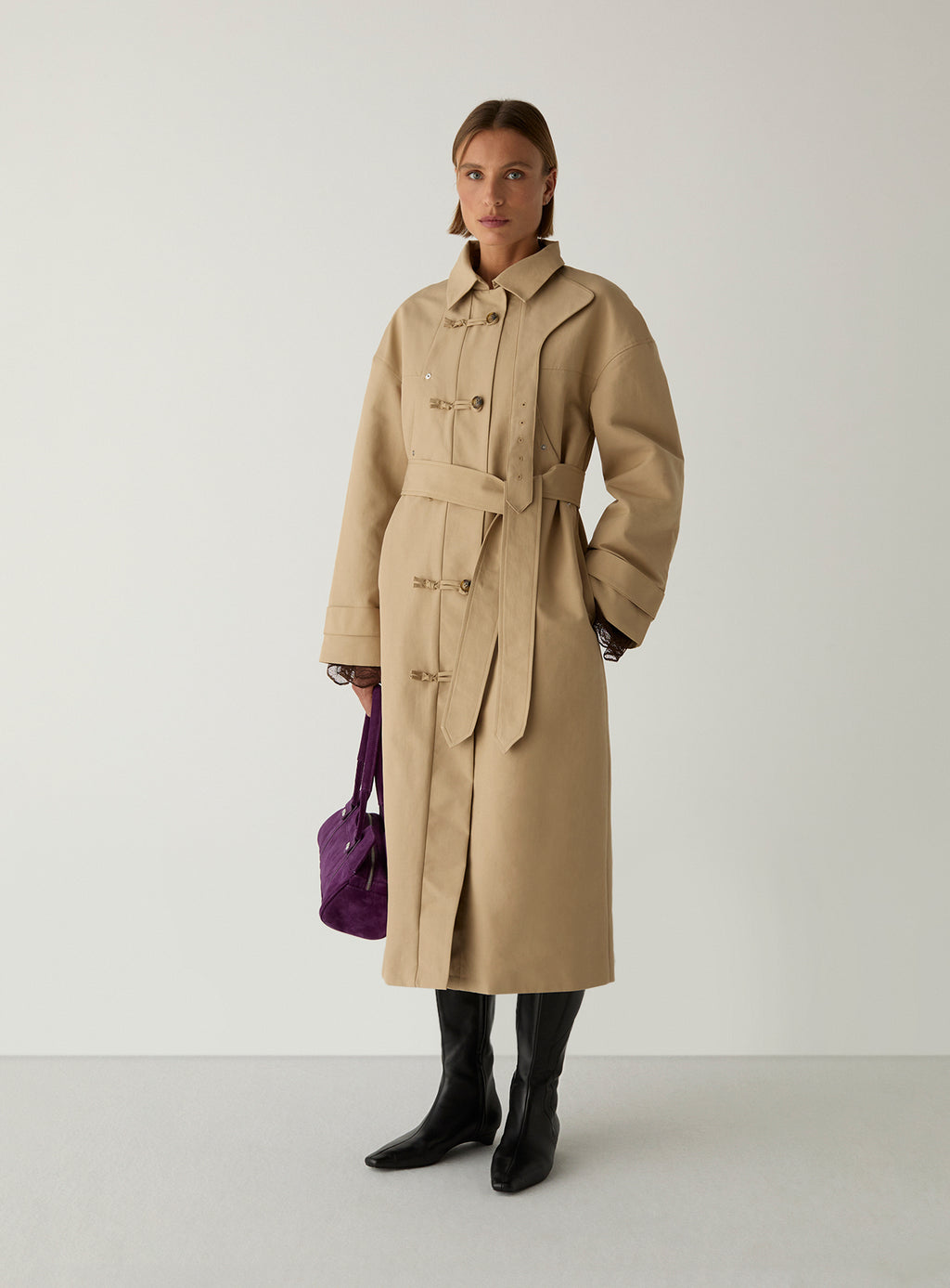 Model wearing the camel Antwerp trench by LAAGAM belted at the waist, with black knee boots and a purple suede bag.