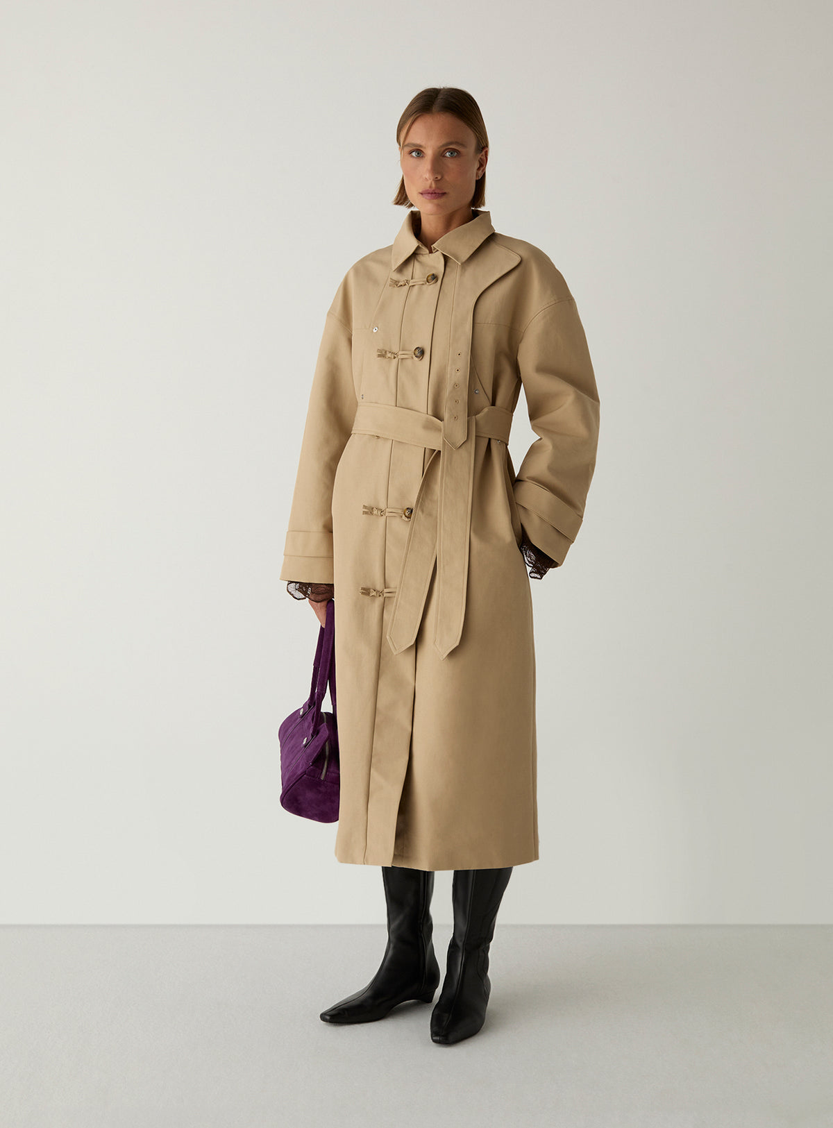 Model wearing the camel Antwerp trench by LAAGAM belted at the waist, with black knee boots and a purple suede bag.