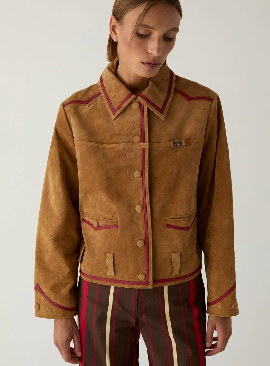 Model wearing the LAAGAM Aveline camel suede jacket with red napa piping, over brown and red striped trousers.